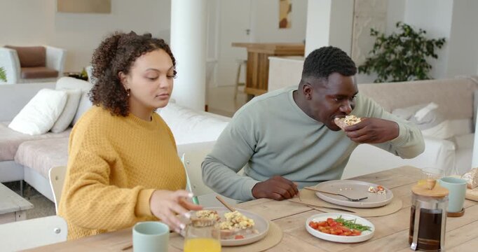 African American couple eating bagel at table, woman offering orange juice after man taking bite