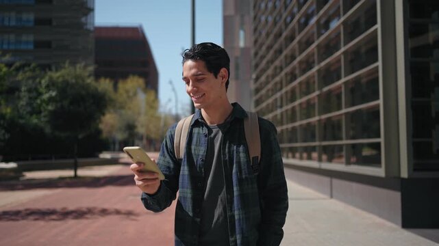 Young student walking outdoors checking smartphone near modern office building in urban city