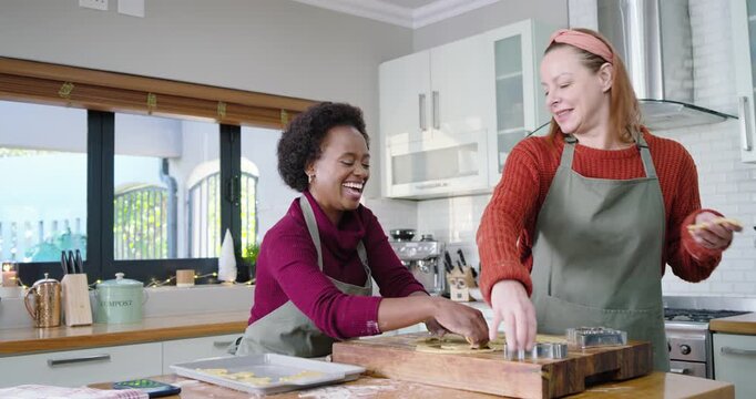 Diverse women smiling, pressing cutters into dough on kitchen island placing shapes for baking