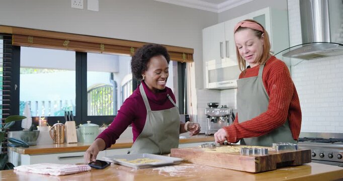 Diverse female friends in aprons laughing at phone, cutting dough, loading oven at counter, baking
