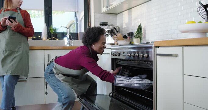 Diverse women friends in aprons taking tray from oven, showing phone, checking, cheering in kitchen