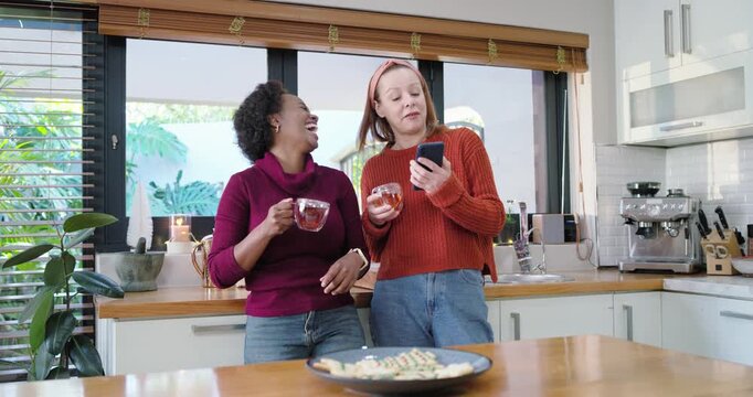Diverse female friends leaning in kitchen holding glass mugs scrolling phone  and  laughing over sna