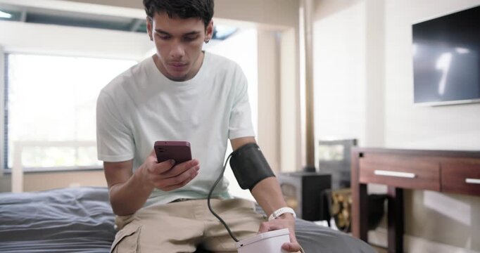 Male adult sitting on bed after cuff getting strapped, holding monitor and checking phone reading