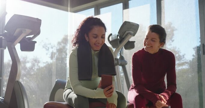 2 female friends on gym bench by windows left scrolling phone sharing spurring right smiling