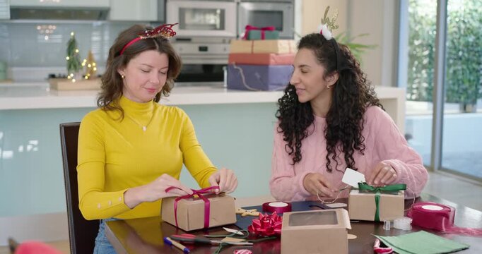 Diverse female friends wrapping kraft gifts at kitchen table for holidays, tying ribbons