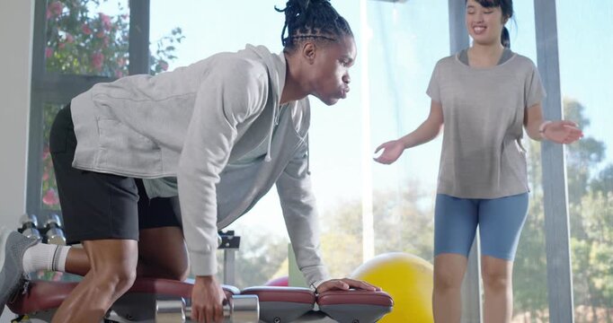 African American man rowing dumbbell at gym bench as Asian coach counting correcting form for power