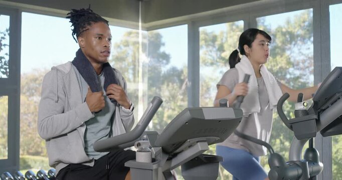 Diverse gym friends warming up for cardio, man pedaling bike, woman using elliptical, towels, water