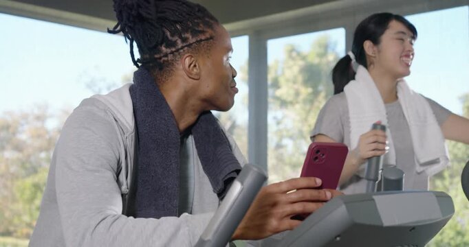 Diverse female friends reacting to magenta phone while left woman tapping console with towel at gym