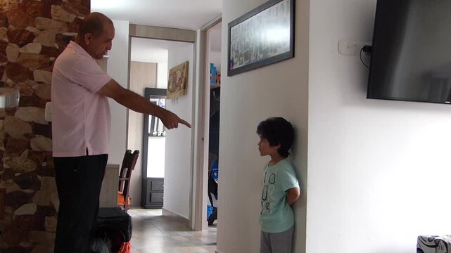 Strict father pointing his finger while scolding or disciplining his guilty looking young son standing against the wall at home