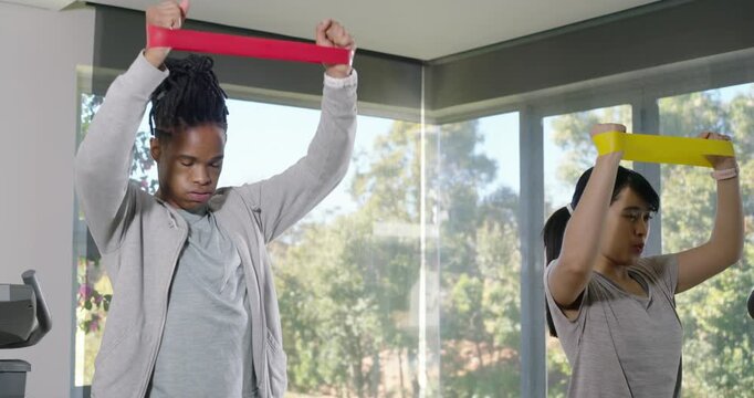 Diverse partners starting, pulling red and yellow bands overhead in sportswear at gym for strength