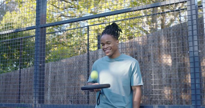 African man wearing teal T-shirt starting balancing neon ball on black paddle, warming up at court