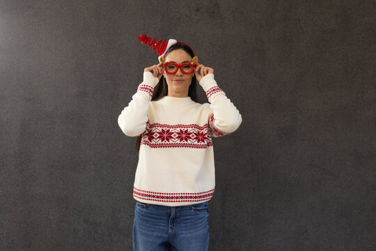 Woman standing and adjusting red novelty glasses against dark grey wall wearing Nordic sweater