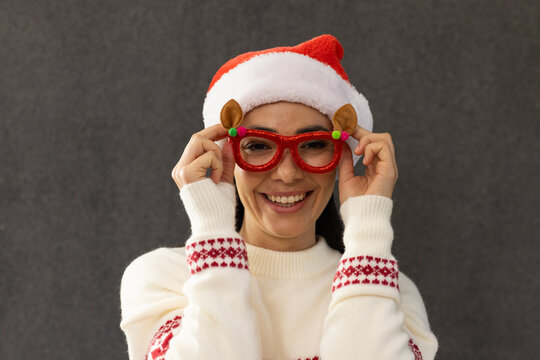 Adult female wearing Santa hat and knit sweater, holding red glitter glasses on dark gray backdrop