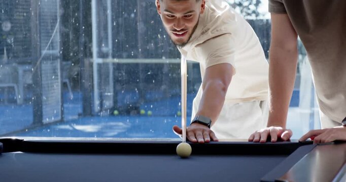 Diverse male friends chatting, shooter leaning over pool table in lounge aiming cue to pocket balls