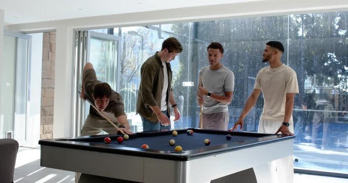 Diverse male friends watching left player lining up and taking shot, playing pool in rec room