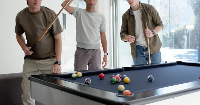 Diverse male friends at home leaning on blue pool table, aiming, striking cue ball, discussing shot