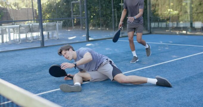 Three men during rally on padel court, gray player lunging, falling, two checking knee with paddles