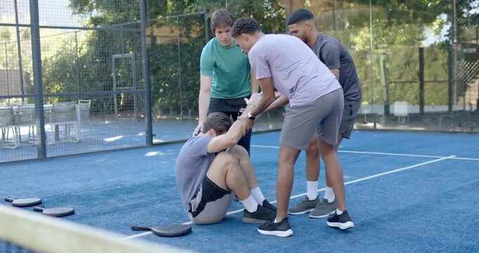 Diverse male team pulling hands, lifting seated player on blue padel court paddles left supporting