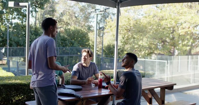 Four males at picnic table, standing male with phone coming left joining chat, pointing at umbrella