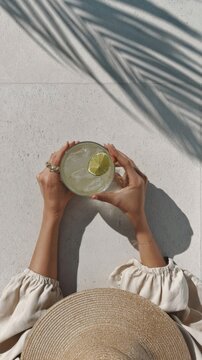 Woman in wide-brim straw hat holding tropical cocktail beside textured white stucco wall with palm leaf shadow, warm sunlight, relaxed resort style, summer fashion portrait