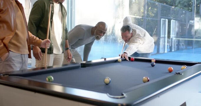 Diverse male friends leaning after shooter lining up and shooting balls on blue-felt table for fun