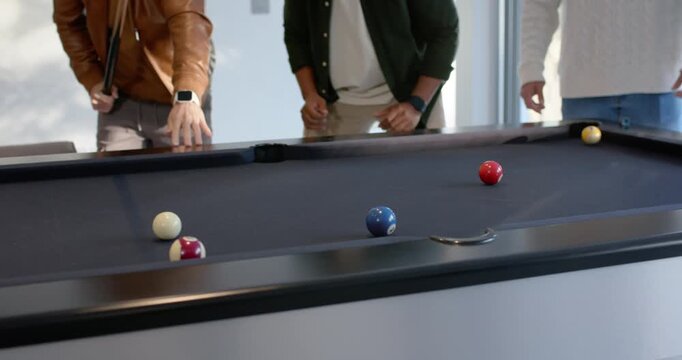 Diverse men watching man aiming striking cue ball at pool table at home pocketing blue ball