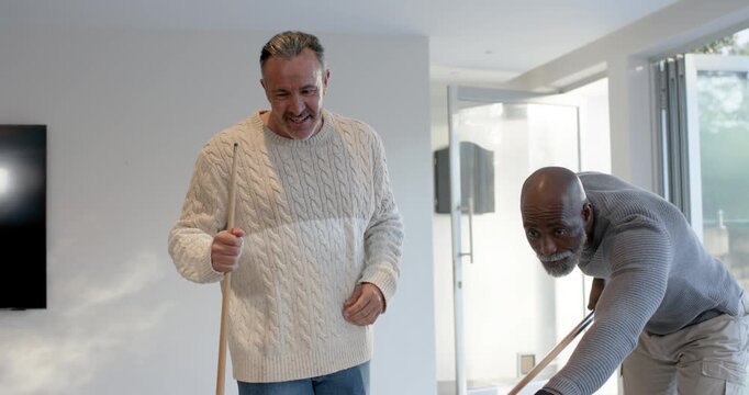 Diverse male friends playing as man in gray sweater lining and striking cue ball, scattering balls