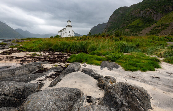 Gimsoy church standing on a rocky sandy beach with mountains and cloudy sky. Lofoten islands, Norway