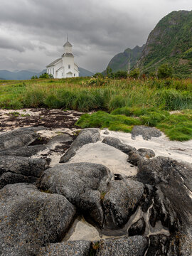 Gimsoy church standing on a grassy hill overlooking a rocky shoreline with mountains under overcast sky. Lofoten Icelands Norway