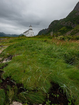 Gimsoy church standing on a grassy hill overlooking a rocky shoreline with mountains under overcast sky. Lofoten Icelands Norway