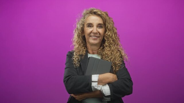 Woman blonde with curly hair holding tablet with arms crossed in studio purple backdrop; friendly confidence.