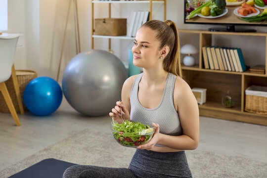 Woman fitness eating salad after home workout. She sits on a yoga mat at home, holding a fresh bowl, exercise balls symbolizing wellness and self care. Clear concept shows healthy lifestyle