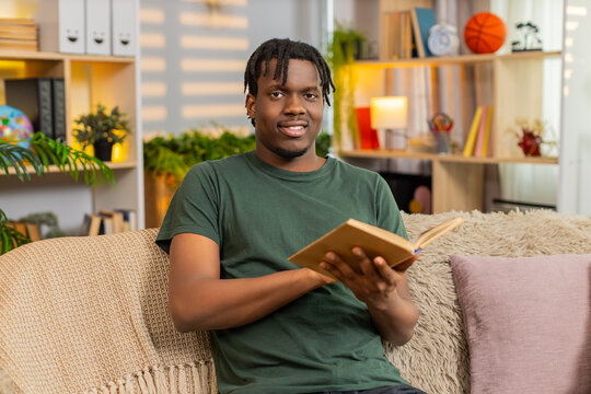 African American young man on home sofa reading open book with curious focus. Black guy on home sofa turning paper page enjoying calm study rest education hobby in warm cozy home living room interior.