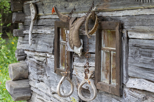Exterior of a wooden log building featuring two small windows, metal chains, wooden rings, and a horned goat skull mounted on the wall during daylight.