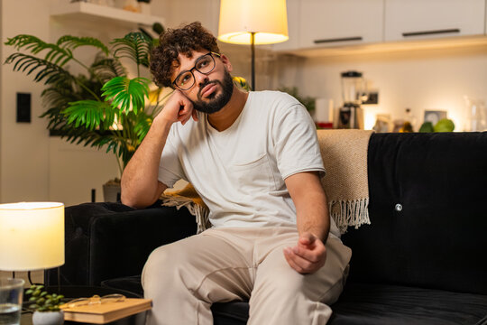 Arabian man at home sits on couch head in hand feels sad after bad news breath slow tired. Egyptian guy on sofa looks down frowns takes deep breath thinks of breakup and hard day seeks calm tonight.