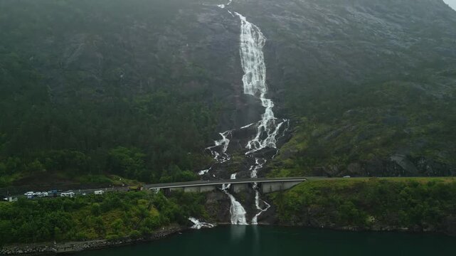 Majestic Langfoss waterfall cascading down rocky cliffs into a serene lake, surrounded by lush greenery and a distant road with parked vehicles