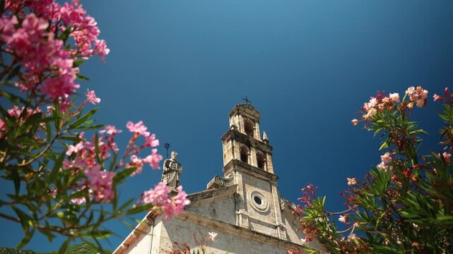 Historic stone church framed by blooming oleander flowers in Prcanj on the Bay of Kotor. Mediterranean architecture under a clear summer sky