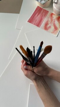 Hand holding assorted paintbrushes beside watercolor artwork, creative hobby, painting tools, artistic practice, visual arts and creative process.