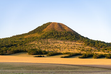 Obraz premium Oblik Hill stands tall in the Bohemian Central Mountains of Czechia. The sun shines on the green slopes and fields nearby. Visitors can enjoy nature and the surrounding landscape.