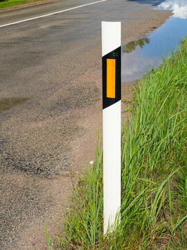White Roadside Delineator Post with Orange Reflector Beside Wet Rural Asphalt Road and Green Grass