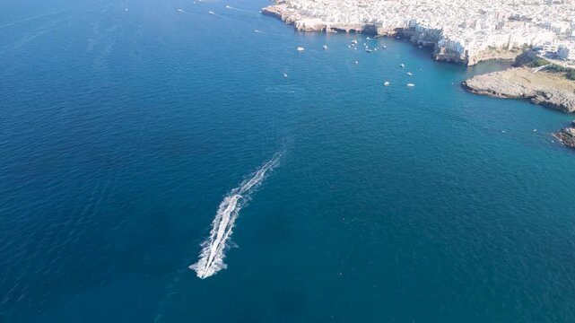 Aerial view of boat wake crossing deep blue sea with white buildings of Polignano a Mare on limestone cliffs above