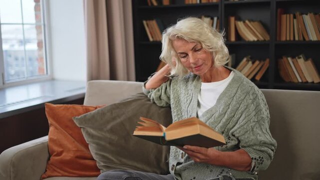 Comfort relaxed senior old elderly woman reading book at home. Mature woman resting enjoy calm day sitting on sofa at home alone. Adult lady spending free time keeping book smiling. Pension concept