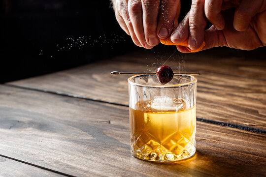 Hands pressing an orange peel releasing the oils on top of an old fashioned cocktail on a wooden bar top.