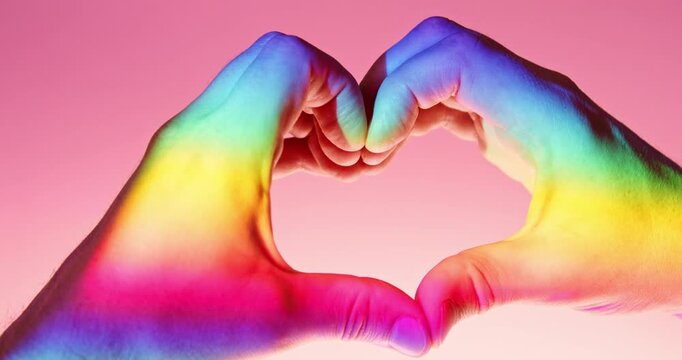 Close-up studio video of two hands forming a heart shape illuminated by rainbow-colored light against a pink background in slow motion. Symbolic LGBTQ+ Pride concept representing love and diversity