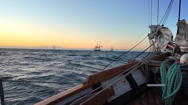 Wide horizontal shot of a wooden sailboat deck pitching heavily on rough sea waves during a golden sunset with distant tall ships on the horizon.