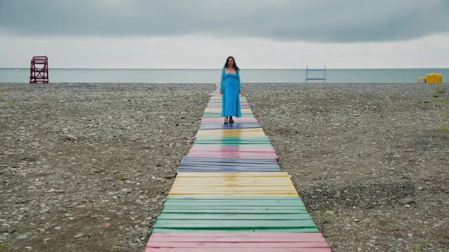 Woman in blue dress walking on rainbow striped wooden path across empty pebble beach toward calm sea on overcast day.