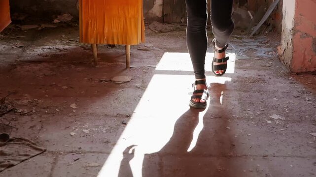 Person wearing black strapped sandals stands on dusty concrete floor with bright sunlight streaming through window opening in deteriorating interior space.