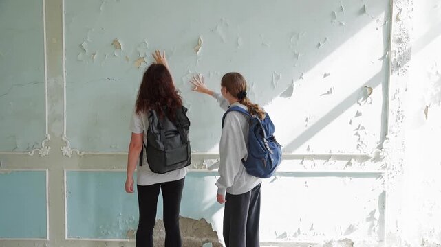 Two young women wearing backpacks stand facing deteriorating wall with chipped paint, sunlight streaming through large window in derelict interior space.