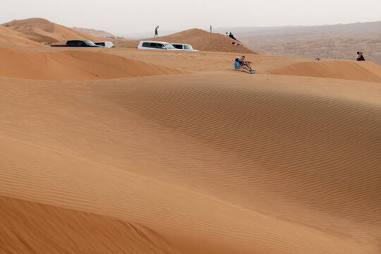 View of Bidiyah sand dunes with people sitting on the crest and off-road vehicles parked nearby in the desert landscape of Bidiyah, Ash Sharqiyah North G, Oman.