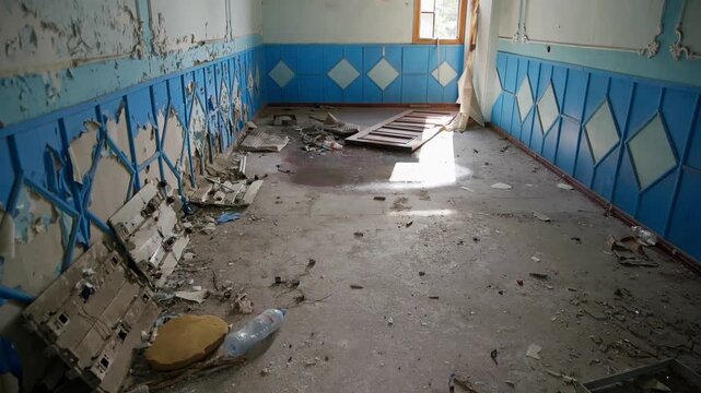 Derelict interior featuring deteriorating blue, cream tile walls, debris scattered on concrete floor, bright window opening casting light into forgotten room.